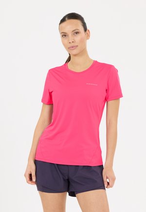Frau mit dunklen Haaren in einem pinkfarbenen kurzärmeligen Sportshirt und marineblauen Shorts steht vor einem weißen Hintergrund.