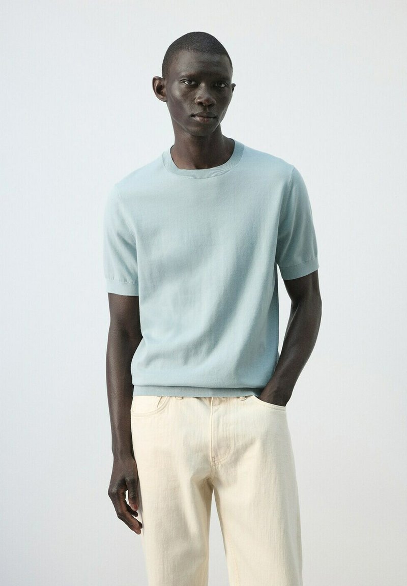 Muž v svetlomodrom svetri s krátkym rukávom a krémových nohaviciach stojí s jednou rukou v vrecku na pozadí hladkej bielej steny.