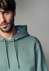 Sudadera verde texturizada con un patrón acolchado, cordones y capucha. El material parece suave y grueso, adecuado para un uso casual.