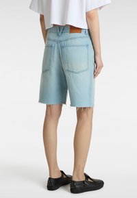 Shorts de mezclilla azul claro con un dobladillo deshilachado, cinco bolsillos y un parche trasero de cuero marrón, combinados con zapatos de lona negros con tachuelas.