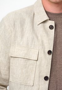 Giacca camicia in lino color beige chiaro con colletto a ribasso, quattro bottoni scuri e taschino sul petto, sovrapposta a un maglione marrone con texture.