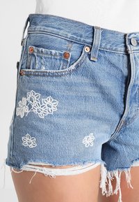 Shorts de denim azul claro con bordado floral blanco, dobladillo deshilachado y remaches de cobre, usados por una persona con muslos visibles y una camiseta blanca.