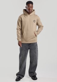 Beige hooded sweatshirt with a front pocket and a circular logo, paired with dark gray wide-leg jeans and black shoes. Smooth texture.