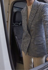 Modrý a bílý tweedový blejzr s dvojitým zapínáním a zlatými knoflíky, nošený přes bílou blůzu, stojící ve dveřích vozidla.