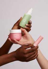 Tre hudpleieprodukter vises: en grønn glassolje-flaske, en rosa kremkrukke og en rosa balsamstift, med minimalistiske hvite etiketter.