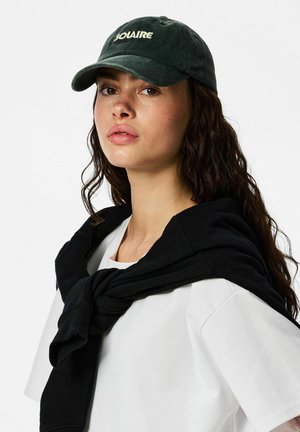 Giovane donna con capelli ricci scuri che indossa un berretto nero con la scritta "SOLAIRE", una maglietta bianca e una felpa nera legata sulle spalle, su sfondo bianco.