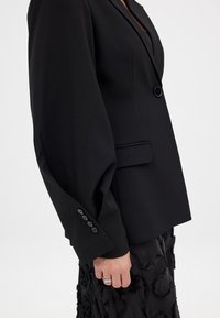 Sort skræddersyet blazer med brede ærmer og en enkelt knaplukning. Har en glat tekstur og detaljerede knapper ved ærmerne.