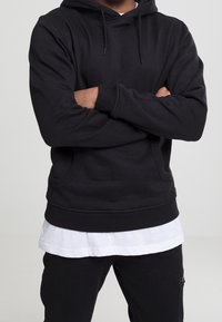 Felpa nera con cappuccio in materiale morbido, con una tasca a marsupio frontale e polsini a costine. Indossata sopra una T-shirt bianca e abbinata a pantaloni scuri.