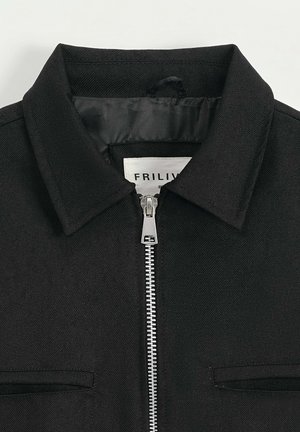 Veste noire en tissu texturé avec un col pointu, une fermeture éclair frontale et deux poches poitrine. L'étiquette indique "FRILIV."