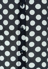 Tessuto nero con grandi pois bianchi, caratterizzato da bottoni blu lungo il centro. Texture liscia con cuciture visibili.