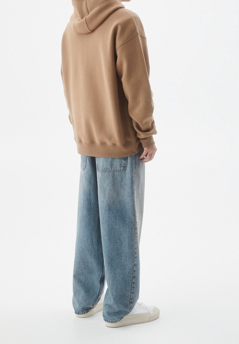 Personne portant un sweat à capuche marron, un jean large bleu clair avec des poches arrière, et des baskets blanches, debout devant un fond blanc uni.