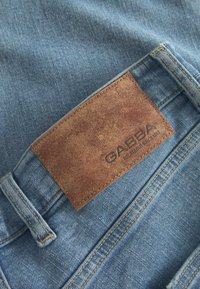 Jeans de denim con una tela de color azul claro, que presentan un parche de cuero marrón desgastado con el logotipo de la marca en relieve, cosido en la parte trasera de la cintura.