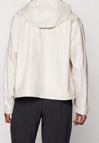 Veste beige clair à capuche avec une coupe courte, tissu texturé, poignets élastiques et détails de surpiqûres contrastantes le long des coutures.