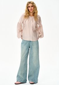 Kobieta z długimi, blond, falującymi włosami, ubrana w jasno-różową bluzkę z falbankami, szerokie jasnoniebieskie dżinsy i różowe buty, stoi na białym tle.