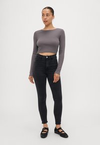 Siv zgornji del s dolgimi rokavi, skrajšan, visoko pasen črn skinny jeans in črne trakaste čevlje. Model stoji na nevtralni podlagi.