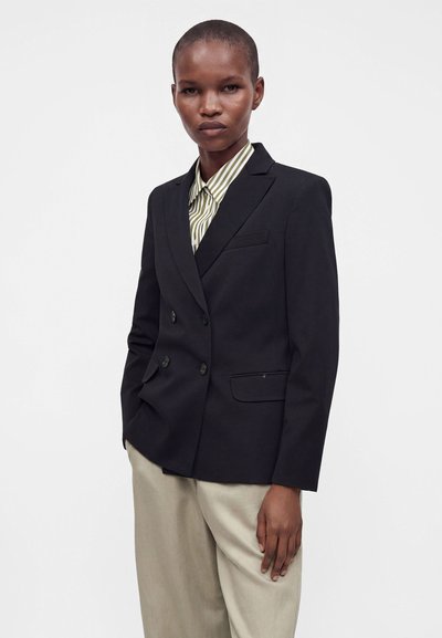 Personne portant un blazer croisé foncé sur une chemise rayée verte et blanche à col, avec un pantalon beige, debout sur un fond blanc.