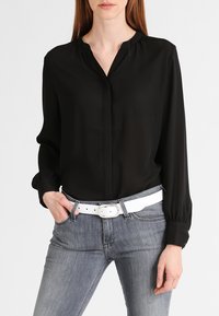 Sort bluse med lange ærmer og krave, der har en glat tekstur. Matchet med grå jeans og et bredt hvidt bælte med en rund spænde.