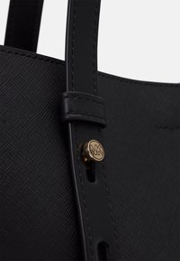 Mustat rakenteelliset kassi, jossa on nahkaiset hihnat, kultainen logo ja ompeluyksityiskohtia. Lähikuva hihnan kiinnityksestä.