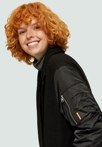 Zwarte jas met een gestructureerd wollen lijf en een glanzende zwarte puffer mouwen. Bevat een zak met rits met een oranje accent op de mouw.