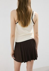 Joven con cabello largo y castaño, vestida con una blusa blanca sin mangas y una falda plisada marrón oscuro, de pie de espaldas contra un fondo liso.