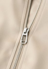 Nahaufnahme eines silbernen Reißverschlusses auf beigem Stoff mit der Gravur "STREET ONE" auf dem Reißverschlussanhänger.