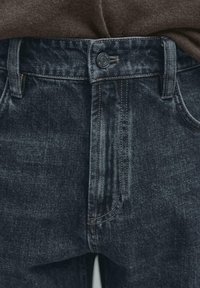 Tmavě prané džíny s hladkou texturou, které mají standardní kapsy, zapínání na knoflík a kontrastní prošívání podél švů.