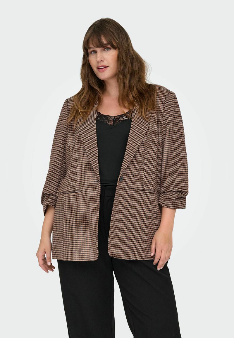 Brūns houndstooth raksts ar pārmērīgu žaketu, ar apakšmalām, garām piedurknēm un sānu kabatām, kombinēts ar melnu topu ar mežģīņu apdari un melnām biksēm.