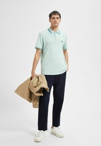 Light blue polo shirt with short sleeves, navy trousers, and white sneakers. Holding a beige jacket. Simple logo on the shirt.