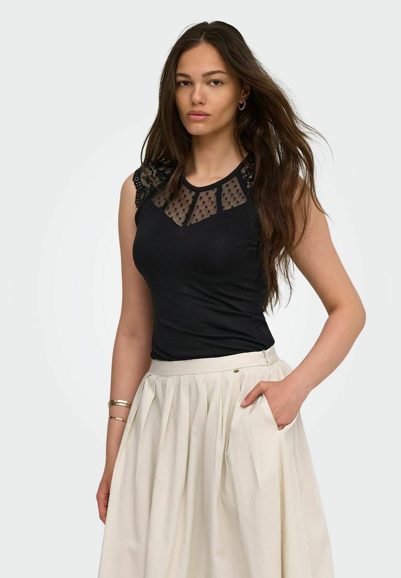 Femme aux longs cheveux foncés portant un haut noir sans manches avec un col en tulle à pois et une jupe plissée blanche, main dans la poche.