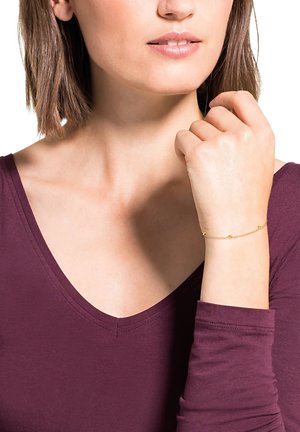 Bracciale d'oro con piccoli accenti rotondi, design a catena. Indossato su un polso, abbinato a un top scollato a V di colore viola scuro. Teint visibile.