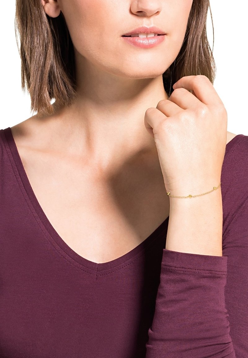 Pulsera de oro con pequeños acentos redondos, diseño de cadena. Llevada en una muñeca, combinada con una blusa de cuello en V morado oscuro. Tono de piel visible.