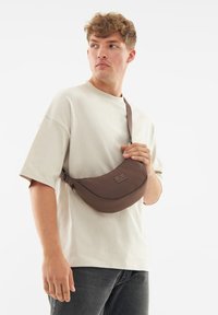 Borsa a tracolla marrone realizzata in tessuto morbido e leggero, con design curvo, tracolla regolabile e etichetta del marchio sul fronte.
