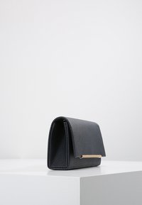 Portefeuille en cuir noir texturé avec un design structuré, comportant un accent en métal doré et une fermeture à rabat. Forme carrée, taille compacte.
