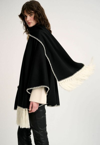 Poncho noir avec franges blanches et broderies décoratives, présentant une coupe ample et un tissu texturé. Porté par-dessus un pull crème et un pantalon noir.