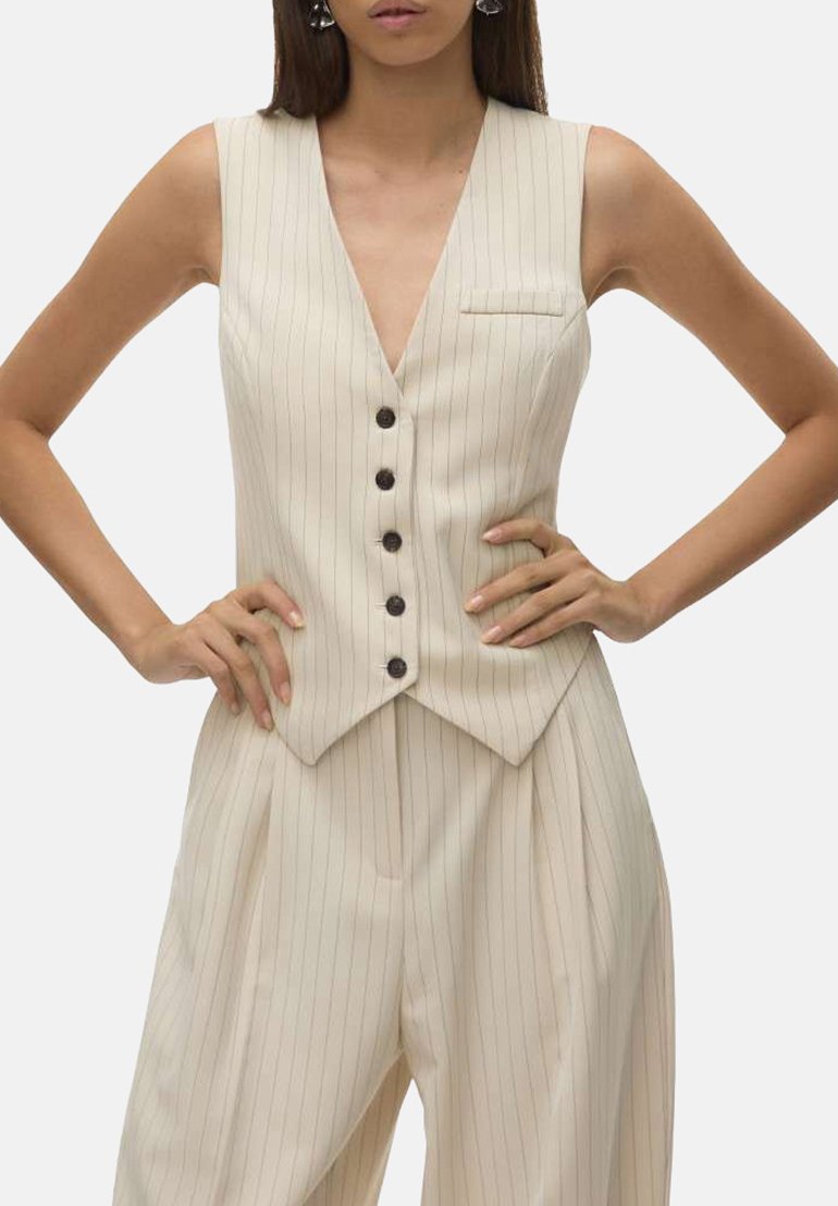 Gilet sans manches de couleur crème, avec un décolleté en V profond, cinq boutons foncés et des rayures verticales. Présente une petite poche poitrine et une forme ajustée.