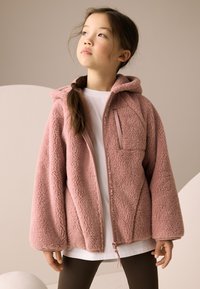 Huppari fleeceä pehmeässä vaaleanpunaisessa sävyssä, jossa on edessä vetoketju, tasku ja leveät hihat. Päällä valkoisen paidan ja ruskeiden ribbineulospolvihousujen kanssa.