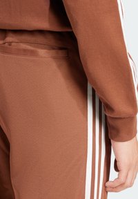 Bruna träningsbyxor med en texturerad tyg, med vita sidostreck och en liten ficka på baksidan.