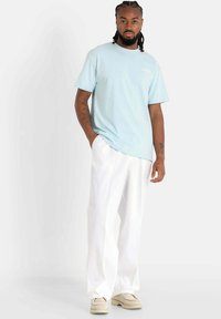 Camiseta de algodón azul claro con mangas cortas, logo blanco en el pecho, combinada con pantalones blancos de corte holgado y zapatos beige.