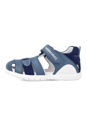 Sandal de cuero azul con diseños troquelados y correa de Velcro. Cuenta con suela de goma blanca y plantilla suave para mayor comodidad. Logotipo de la marca visible.