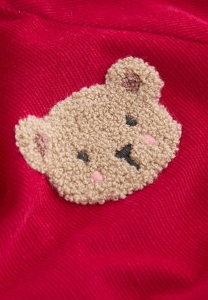 Broderad björnsans patch i beige med svarta ögon och en rosa näsa på en röd texturerad tygbakgrund.