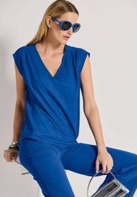 Blaues ärmelloses Oberteil mit V-Ausschnitt, kombiniert mit passenden blauen Hosen. Das Modell hält eine silberne Handtasche. Der Look zeichnet sich durch glatte, elegante Stoffe aus.