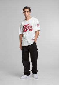 Maillot blanc à manches courtes avec lettrage et accents rouges, présentant un devant à boutons. Associé à un pantalon cargo noir et des baskets blanches.