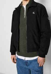 Veste bomber légère noire avec poignets élastiques, présentant un détail de logo subtil. Portée sur un pull côtelé vert et une chemise blanche.