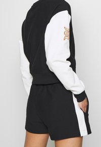 Felpa bianca e nera con design cropped e logo ricamato arancione sulla manica, abbinata a pantaloni corti neri coordinati.
