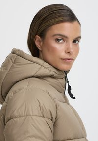 Beige puffer-jakk, millel on kapuuts, jaotatud tikandiga ning musta nööriga. Sile tekstuur ja keha lähedane lõige, kantud mudeli poolt.
