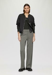 Schwarze Bomberjacke mit rundem Kragen, graue lässige Hose, klobige schwarze Schuhe und eine kleine schwarze Umhängetasche.