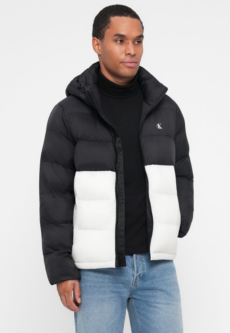 Gesteppten Jacke mit der oberen Hälfte in Schwarz und der unteren Hälfte in Weiß, ausgestattet mit einem Reißverschluss, Kapuze und einem Logo auf der Brust. Material aus Baumwollmischung.