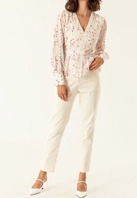 Blusa bianca a maniche lunghe con motivo floreale rosso, rifinitura in pizzo e vita a peplo. Abbinata a pantaloni beige su misura e tacchi bianchi.