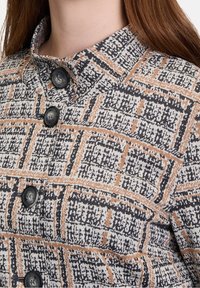 Skjorta med knappar i ett vävt mönster som har svarta, vita och orangea nyanser. Utmärkande krage och textil med stor svart knappar.