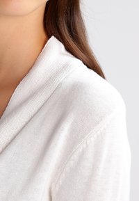 Suéter blanco de punto con cuello tipo chal, que presenta una textura fina y sutilezas en los detalles de costura en los hombros.